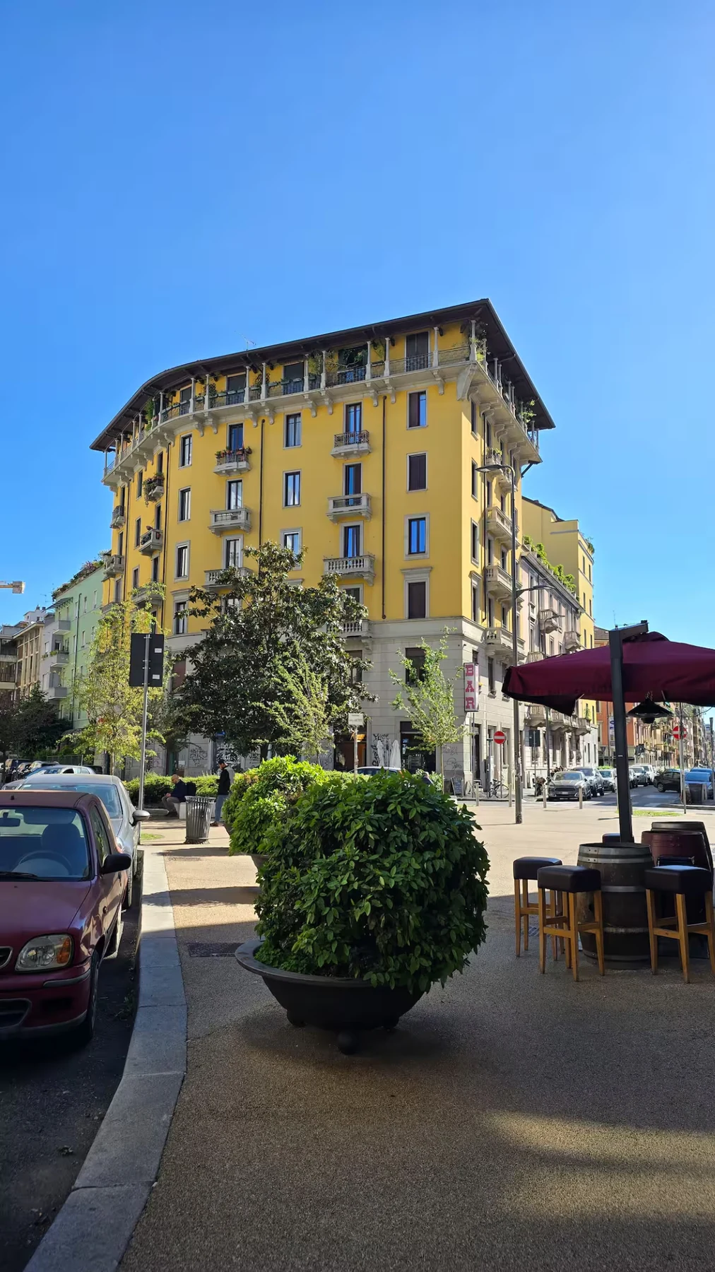 Street-level NoLo corner with the yellow Milan building, café seating and trees.