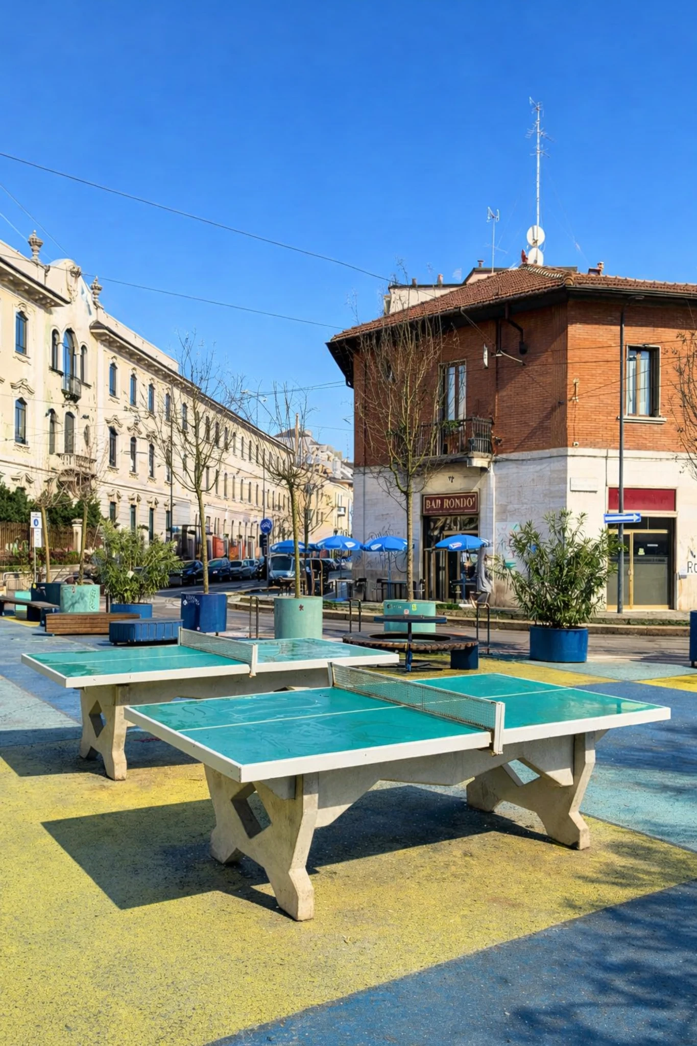 Colorful public space in NoLo with outdoor ping-pong tables, surrounding buildings and clear blue sky.